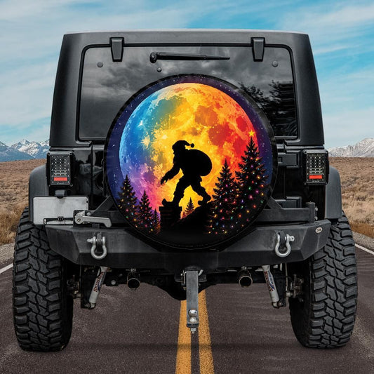 A circular spare tire cover mounted on the rear tire of a black Jeep, featuring a colorful rainbow moon and a black silhouette of a Bigfoot figure walking through pine trees. The Jeep is shown from the back on an asphalt road with mountains and a blue sky in the background.